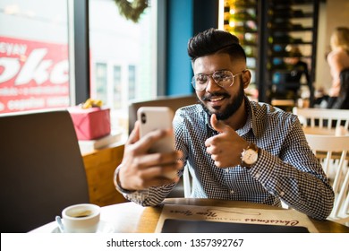 Young Indian Man Sitting At Cafe Making Video Call From His Mobile Phone. Asian Male At Coffee Shop Having A Videochat On Smart Phone.