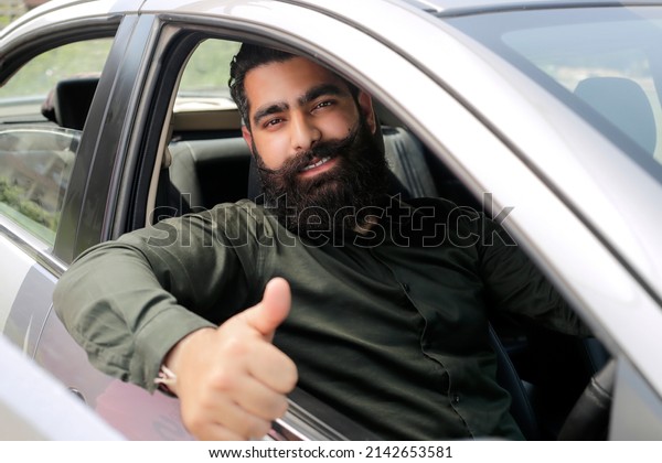 Young Indian man is sitting in car and showing\
thumbs up sign.