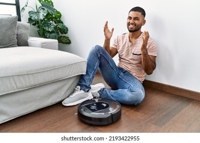 Young Indian Man Sitting At Home By Vacuum Robot Celebrating Victory With Happy Smile And Winner Expression With Raised Hands 