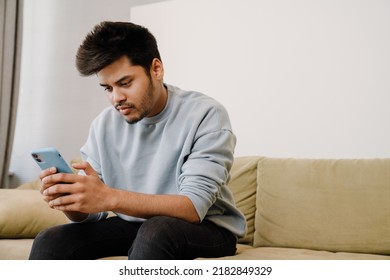 Young Indian Man Sitting On Sofa And Scrolling News On His Phone At Home