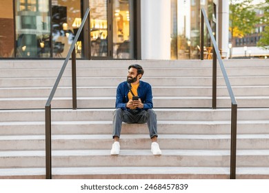 A young Indian man is sitting on the steps of a modern outdoor staircase in an urban area. He appears relaxed, holding a smartphone, and looking off into the distance. - Powered by Shutterstock