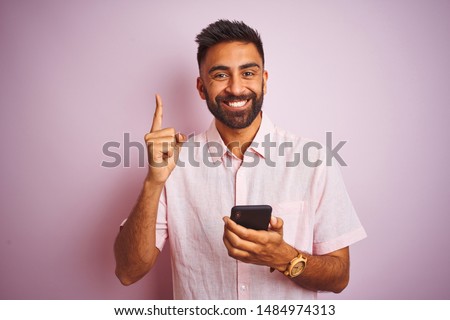 Young indian man using smartphone standing over isolated pink background surprised with an idea or question pointing finger with happy face, number one