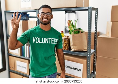 Young Indian Man Volunteer Holding Donations Box Showing And Pointing Up With Fingers Number Three While Smiling Confident And Happy. 