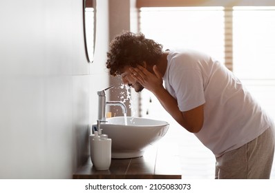 Young indian man washing face with clear water from tap bending over sink, male model doing cleanup skincare facial routine in bathroom in the morning, side view - Powered by Shutterstock