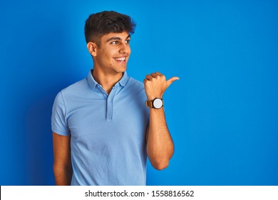 Young Indian Man Wearing Casual Polo Standing Over Isolated Blue Background Smiling With Happy Face Looking And Pointing To The Side With Thumb Up.