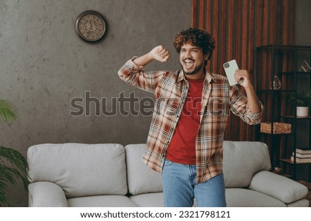 Young Indian man wearing casual clothes hold in hand use mobile cell phone do winner gesture stand near grey sofa couch stay at home hotel flat rest relax spend free spare time in living room indoor