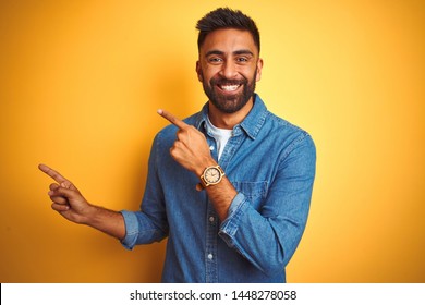 Young indian man wearing denim shirt standing over isolated yellow background smiling and looking at the camera pointing with two hands and fingers to the side.