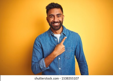 Young Indian Man Wearing Denim Shirt Standing Over Isolated Yellow Background Cheerful With A Smile Of Face Pointing With Hand And Finger Up To The Side With Happy And Natural Expression On Face