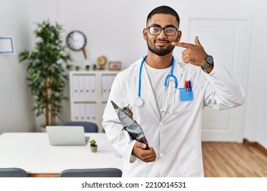 Young Indian Man Wearing Doctor Uniform And Stethoscope Pointing With Hand Finger To Face And Nose, Smiling Cheerful. Beauty Concept 