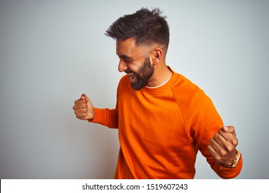 Young Indian Man Wearing Orange Sweater Over Isolated White Background Very Happy And Excited Doing Winner Gesture With Arms Raised, Smiling And Screaming For Success. Celebration Concept.