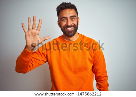 Young indian man wearing orange sweater over isolated white background showing and pointing up with fingers number five while smiling confident and happy.