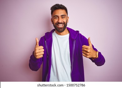 Young indian man wearing purple sweatshirt standing over isolated pink background success sign doing positive gesture with hand, thumbs up smiling and happy. Cheerful expression and winner gesture. - Powered by Shutterstock