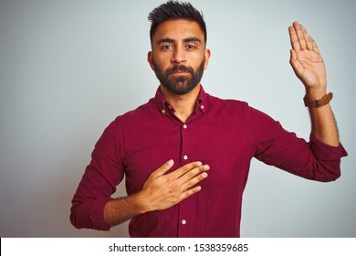 Young indian man wearing red elegant shirt standing over isolated grey background Swearing with hand on chest and open palm, making a loyalty promise oath