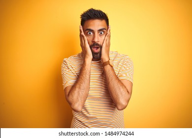 Young indian man wearing t-shirt standing over isolated yellow background afraid and shocked, surprise and amazed expression with hands on face