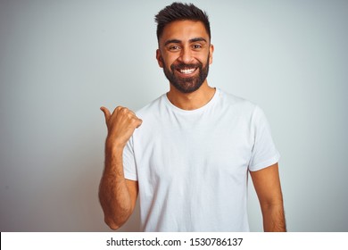 Young Indian Man Wearing T-shirt Standing Over Isolated White Background Smiling With Happy Face Looking And Pointing To The Side With Thumb Up.