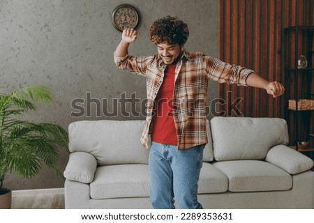 Young Indian man wears casual clothes dancing have fun gesticultating hands stand near grey sofa couch stay at home hotel flat rest relax spend free spare time in living room indoor. Lounge concept