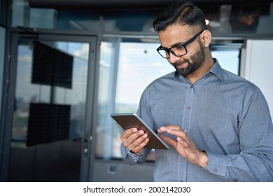 Young indian marketing business manager expert in glasses standing in modern corporate office using digital tablet device searching analyzing project results. Business technologies concept. - Powered by Shutterstock