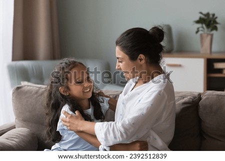 Young Indian mother playing with little preschooler daughter seated on sofa at home, laughing, hugging feel happy, enjoy reunion after separation. Unconditional love, cherish, family bond, fun concept