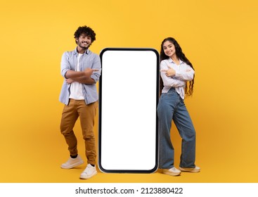 Young Indian Spouses Posing Near Huge Smartphone With Mockup, Promoting Mobile App Or Website, Advertising Your Service Or Product Over Yellow Studio Background, Free Space