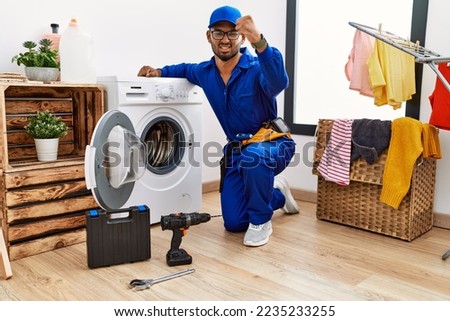 Young indian technician working on washing machine angry and mad raising fist frustrated and furious while shouting with anger. rage and aggressive concept. 