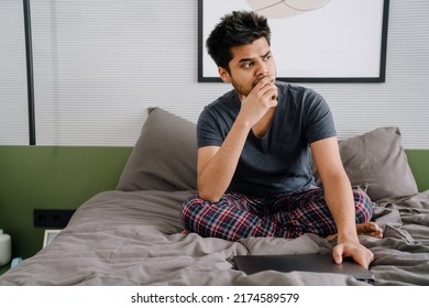 Young Indian Thoughtful Man Touching Chin With His Hand Holding Closed Laptop Looking Rightward Sitting On Bed At Home