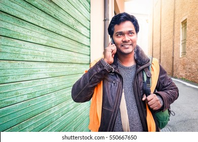 Young Indian Travel Man Making Phone Call Walking On The Street Holding Bag At Winter Time - Handsome Bangladeshi Guy Looking Up Using Mobile Smiling Outdoors  - Communication Concept With Copy Space