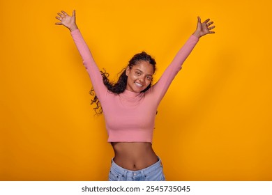 Young Indian woman beams with happiness, her arms stretched wide in celebration. She wears a fitted pink top and casual jeans, radiating positivity against a bright yellow background. - Powered by Shutterstock