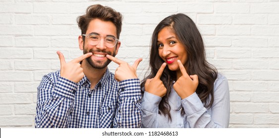 Young indian woman and caucasian man couple smiles, pointing mouth, concept of perfect teeth, white teeth, has a cheerful and jovial attitude