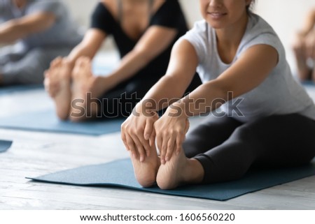 Young Indian woman doing Seated forward bend exercise, feet close up, diverse people practicing yoga at group lesson, stretching in paschimottanasana pose on mat, working out in modern yoga studio