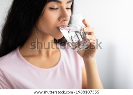 Young Indian woman drinking clear mineral water from glass at home, selective focus. Lovely Asian lady staying hydrated, taking care of her body. Healthy living and detox concept