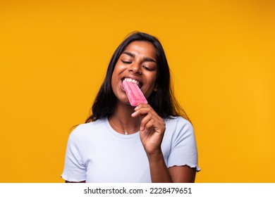Young Indian woman eating pink popsicle on yellow background. Asian woman eating ice cream.