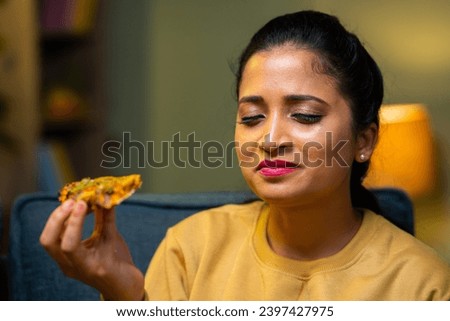 Young Indian woman enjoying byte of pizza at home during evening - concept of satisfied, foodie and delightful expression