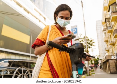 Young Indian Woman In Face Mask Using Cellphone While Standing With Powered Scooter Outdoors