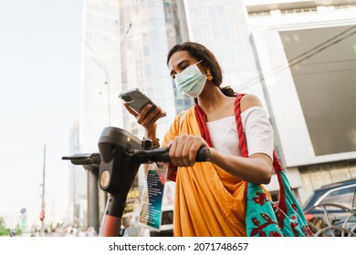 Young Indian Woman In Face Mask Using Cellphone While Standing With Powered Scooter Outdoors