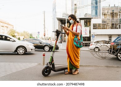 Young Indian Woman In Face Mask Using Cellphone While Standing With Powered Scooter Outdoors