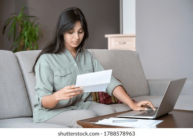 Young indian woman holding letter using laptop computer application paying bill online on website, managing account finances, calculating budget tax banking loan debt payment sitting on couch at home.