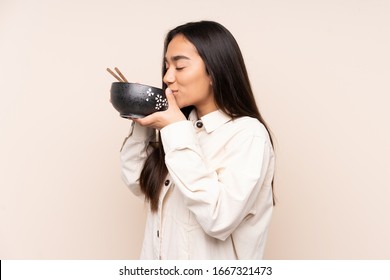Young Indian woman isolated on beige background holding a bowl of noodles with chopsticks and eating it