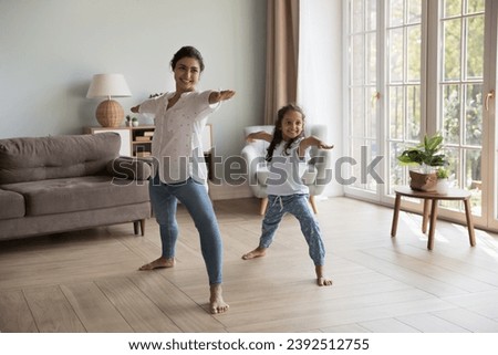Young Indian woman and little girl perform asanas at modern home. Mom teach daughter yoga do together Warrior one exercise, standing barefoot in cozy warm living room. Healthy lifestyle, sport concept