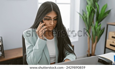 A young indian woman with long hair works at her office desk while talking on a smartphone, embodying professionalism and concentration.
