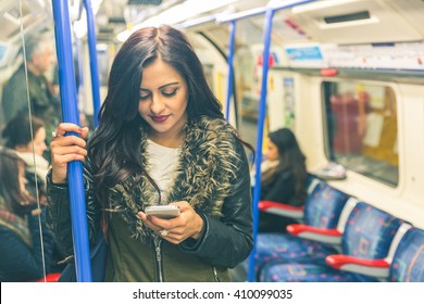 Young Indian Woman Looking At Her Smart Phone And Typing While Travelling On The Tube In London. She Is Standing And Holding With An Hand. Transport And Commuting Concepts. Colours Filter Added.