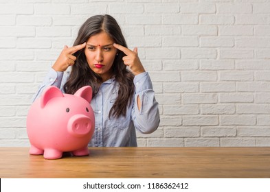 Young Indian Woman Man Making A Concentration Gesture, Looking Straight Ahead Focused On A Goal. Holding A Piggy Bank.