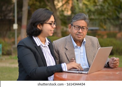 Young Indian Woman Manager/entrepreneur In Western Formals Or Suit Helping Old Indian Man On A Laptop Promoting Digital Literacy For Elderly In A Park In New Delhi, India