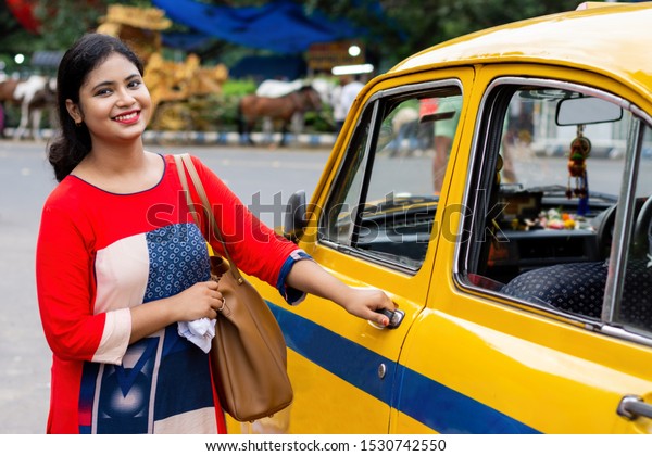 Young\
Indian woman opening gate of old yellow taxi\
