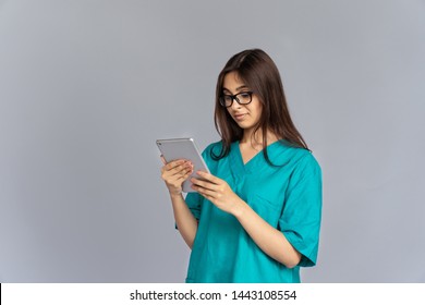 Young Indian Woman Professional Nurse Female Doctor Wear Green Uniform Glasses Holding Using Digital Tablet Consult Patient In Online Chat Video Calling Isolated On Grey Studio Background, Copy Space