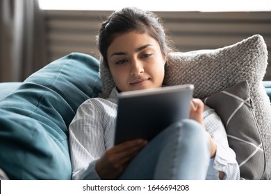 Young Indian woman relax on cozy sofa furniture in living room using browsing Internet on tablet, millennial ethnic girl lying resting on comfortable couch at home watching movie or video on pad - Powered by Shutterstock