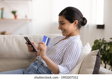 Young indian woman shopper sit on couch enjoy easy paying for goods online using bank card phone. Happy mixed race female traveler book tickets trip hotel at travel agency transport operator website