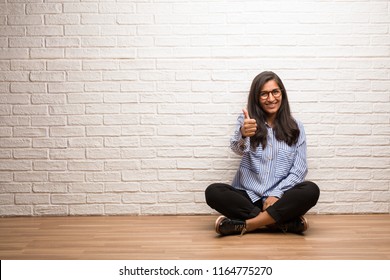 Young indian woman sit against a brick wall cheerful and excited, smiling and raising her thumb up, concept of success and approval, ok gesture - Powered by Shutterstock