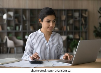 Young Indian woman sit at desk calculate expenses expenditures on calculator pay bills taxes online on laptop. Millennial female manage household budget make payment on computer use internet.