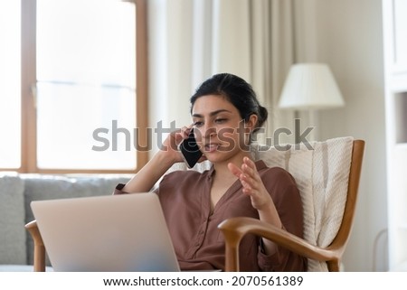 Young Indian woman sit on armchair looks at laptop screen while talks on phone, solve business by phonecall. Mobile carrier operator, modern tech usage, remote conversation, make order online concept