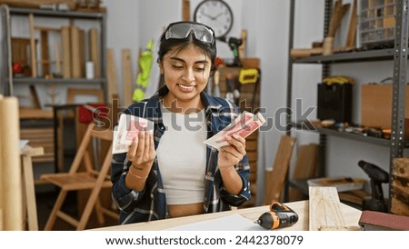 A young indian woman smiles while holding icelandic krona in a well-organized carpentry workshop.